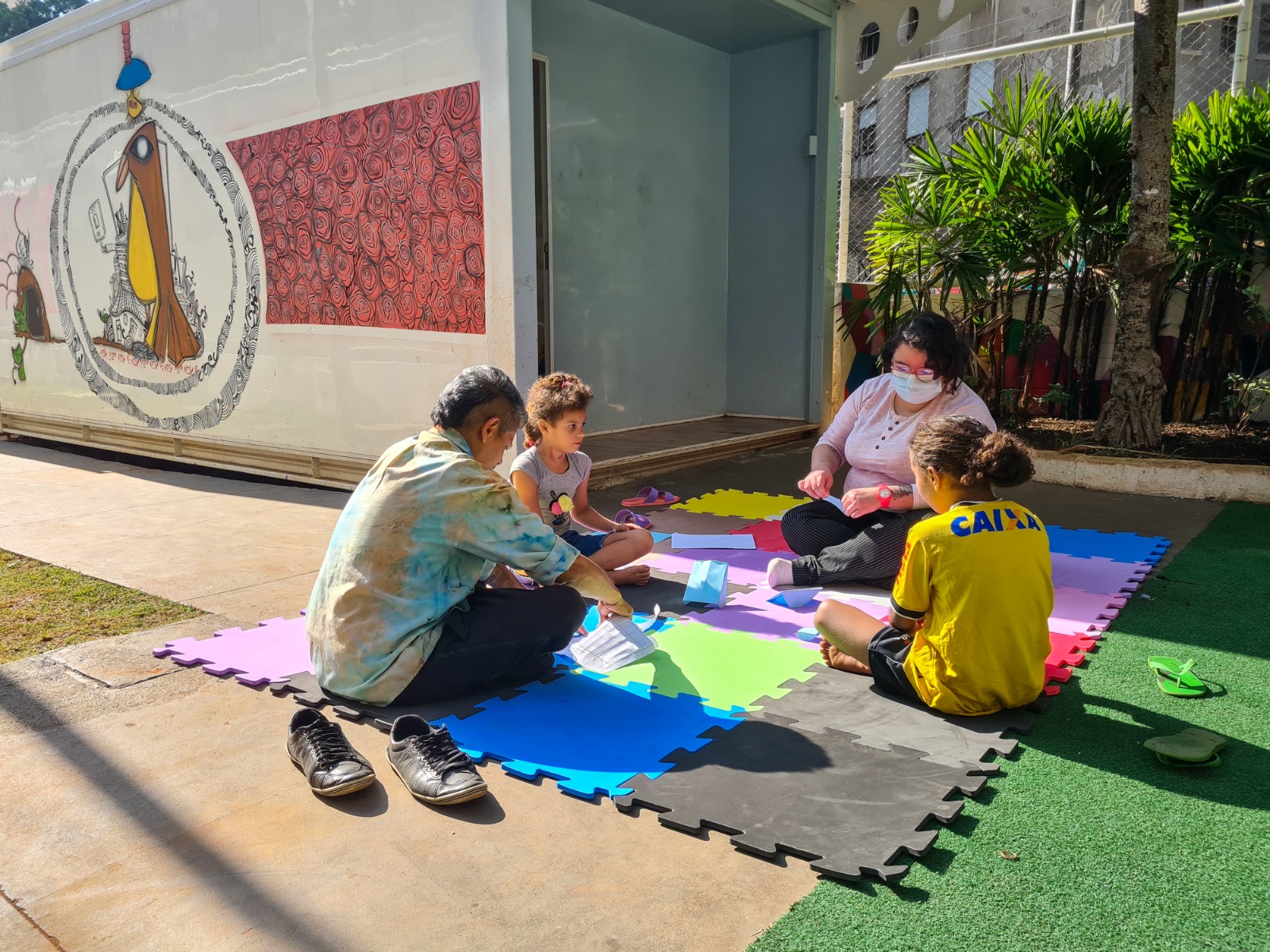 2 crianças e 2 mulher se sentam no chão sob um tapete colorido. Eles estão com papéis coloridos em mãos e em sua frente, fazendo dobraduras. No fundo há árvores e uma casa branca, a qual tem uma parede com grafite colorido.